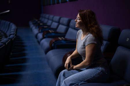Caucasian red-haired woman sits on the last row in a cinema in an empty hall. The girl is watching a movie alone.の写真素材