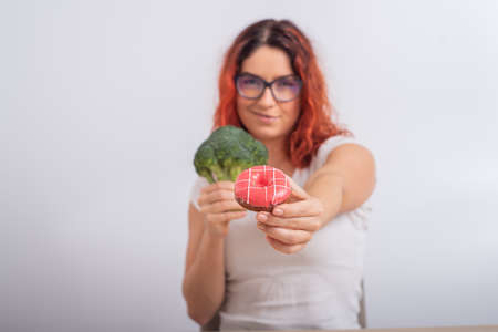 Caucasian woman chooses between vegetables and fast food. Redhead girl holding broccoli and donut on a white background.の写真素材