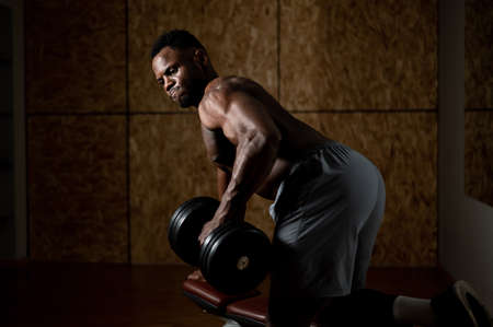 African american man with naked torso doing triceps row with dumbbell on bench.の写真素材