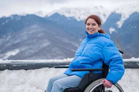 A happy woman dressed in a blue coat sits in a wheelchair on a point view and looking at the snow-capped mountains.の写真素材