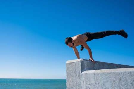 Shirtless man doing a handstand on a parapet by the sea.