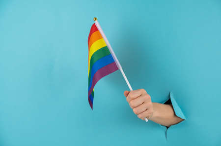A womans hand sticks out of a hole in a blue paper background and holds an lgbt flag.の写真素材