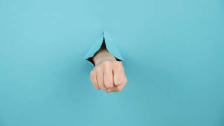 A womans hand sticking out of a hole from a blue background shows a fist.の写真素材