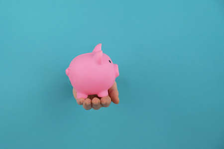 A female hand sticking out of a hole from a blue background holds a pink piggy bank.の写真素材