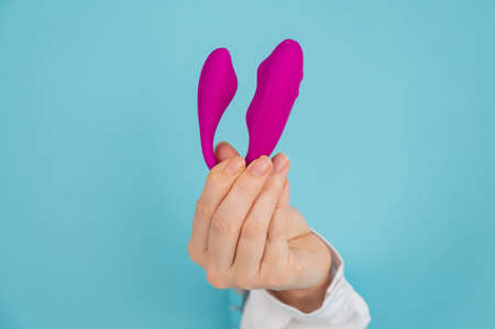A womans hand holds a pink sex toy through a hole in a paper blue background.