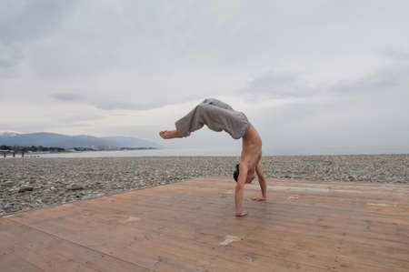 Shirtless caucasian man doing backflip on pebble beach.