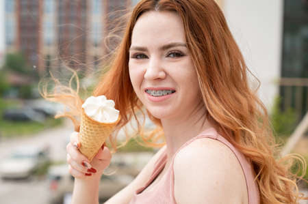 Portrait of young beautiful red-haired woman smiling with braces and going to eat ice cream cone outdoors in summerの写真素材