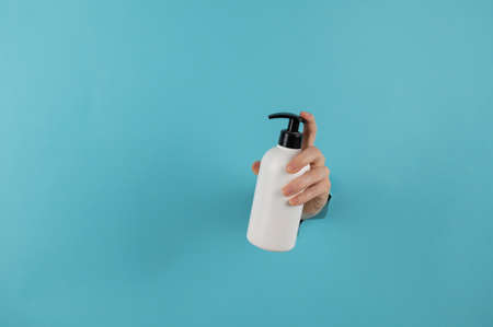 A womans hand sticks out of a hole in a blue paper background and holds a skin lotion pump.の写真素材