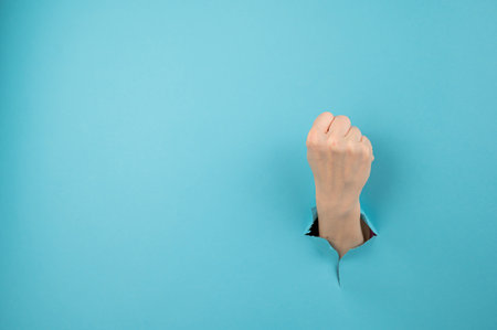 A womans hand sticking out of a hole from a blue background shows a fist.の写真素材