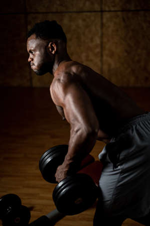 African american man with naked torso doing triceps row with dumbbell on bench.の写真素材
