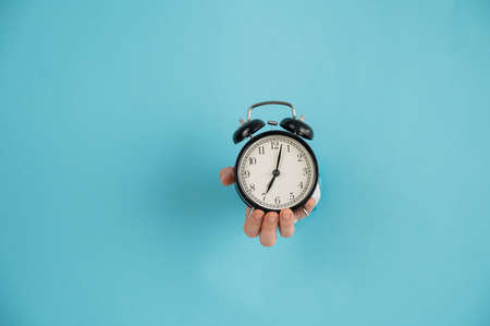 A womans hand sticks out of a hole in a blue paper background and holds an alarm clock. Copy space.
