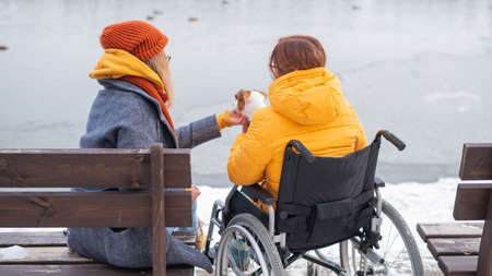 Caucasian woman in a wheelchair and her friend are sitting by the lake with ducks in winter.の写真素材