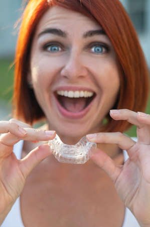 Red-haired Caucasian woman holding transparent mouthguards for bite correction outdoors. A girl with...