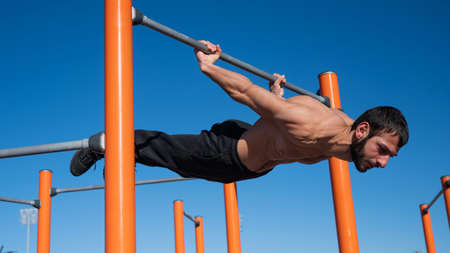 Shirtless man doing blanche on horizontal bar outdoors.