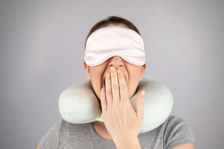 Caucasian woman yawns with travel pillow and sleep mask on white background.