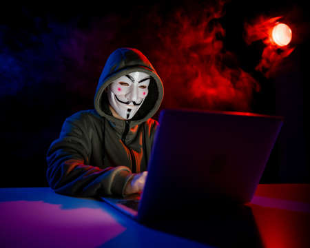 June 5, 2022 Novosibirsk, Russia: Anonymous in a hood is typing on a laptop in the dark in red-blue...
