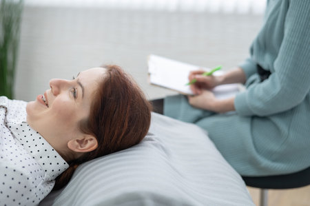 A caucasian woman lies on a couch and expresses her feelings, while a psychologist makes notes on a tablet.の写真素材
