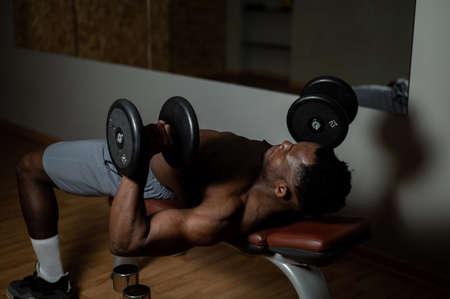 Shirtless afro american man doing exercises with dumbbells lying on bench in gym.