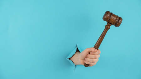 A womans hand with a wooden judges gavel sticks out of a hole in a blue background. Copy space.