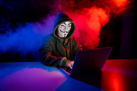 June 5, 2022 Novosibirsk, Russia: Anonymous in a hood is typing on a laptop in the dark in red-blue smoke.のeditorial素材