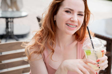 Young beautiful red-haired woman with braces drinks cooling lemonade outdoors in summer. Portrait of...