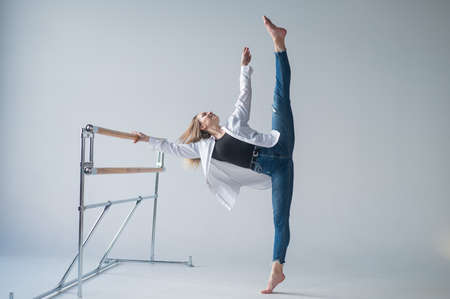 Caucasian woman in casual clothes pulls the split at the ballet barre.