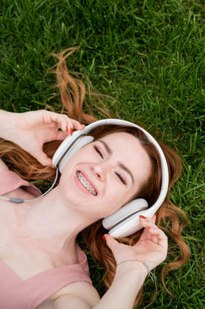A young red-haired woman lies on the grass and listens to music.
