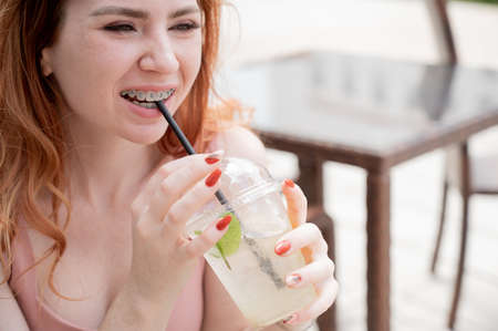 Young beautiful red-haired woman with braces drinks cooling lemonade outdoors in summer. Portrait of a smiling girl with freckles.の写真素材