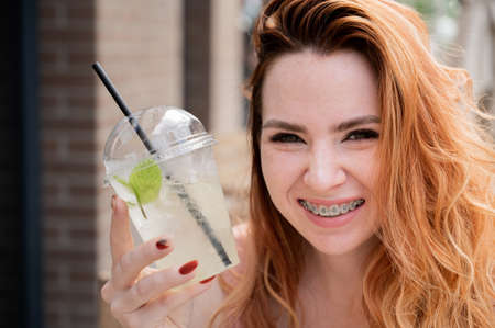 Young beautiful red-haired woman with braces drinks cooling lemonade outdoors in summer. Portrait of a smiling girl with freckles.の写真素材