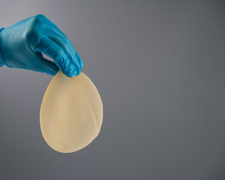 A female hand in gloves holds a teardrop-shaped breast implant on a gray background.の写真素材