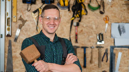 Portrait of a carpenter in goggles and overalls holding a wooden hammer in the workshop against the background of a wall with tools.の写真素材