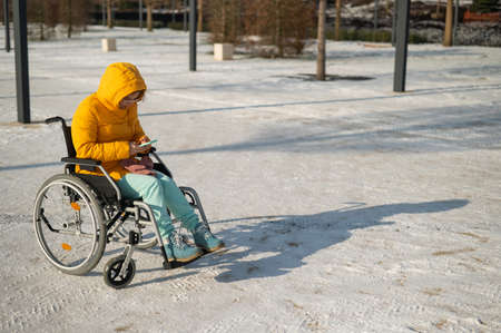 Caucasian woman in a wheelchair uses a smartphone on a walk in the park in winter.