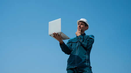 A male builder in a helmet stands against a blue sky with a laptop in his hands.