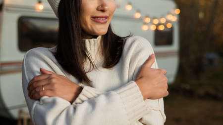A pensive Caucasian woman in a white knitted sweater and a hat is resting near a motorhome in a warm autumn. Travel by camper.の写真素材
