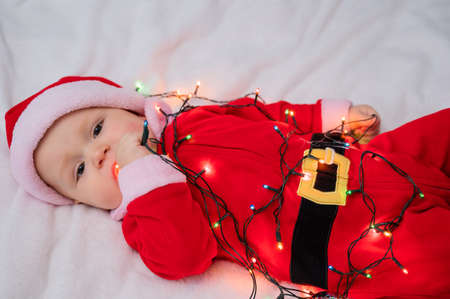 A baby boy dressed in a santa suit lies on the bed and plays with a Christmas garland.