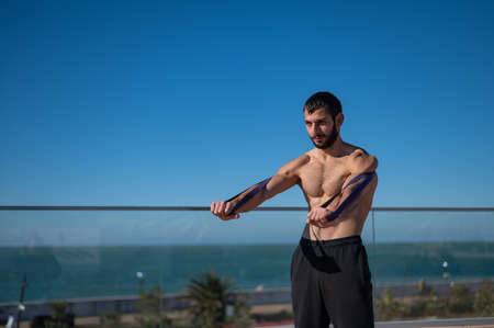 Shirtless man doing exercise with rubber bands outdoors.