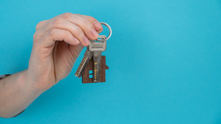 A womans hand holds the keys with a keychain in the shape of a house on a blue background.の写真素材