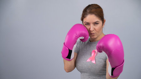 Caucasian woman in pink boxing gloves with a pink ribbon on her chest on a gray background. Fight...