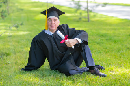 An elderly gray-haired man in a graduation gown sits on green grass.の写真素材