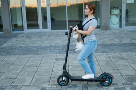 A woman rides an electric scooter with a dog in a backpack. Pappilion Spaniel Continental in a...