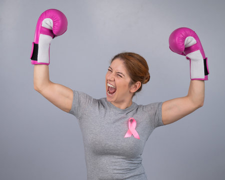 A woman with a pink ribbon on her chest raises her hands in pink boxing gloves on a gray background. Victory over breast cancer.の写真素材
