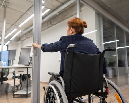 Red-haired caucasian woman in a wheelchair trying to open the door in the office.の写真素材