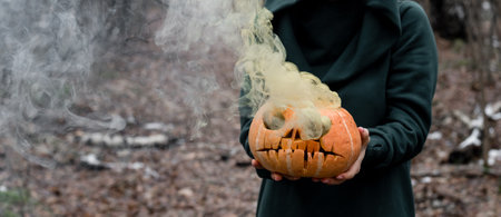 A creepy witch holds a steaming pumpkin in a deep forest. Jack o lantern emits yellow smoke for...