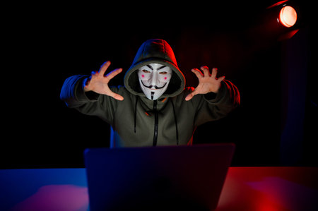 June 5, 2022 Novosibirsk, Russia: Anonymous in a hood is typing on a laptop in the dark in red blue...