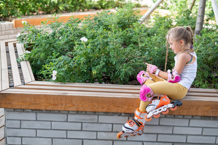 A little Caucasian girl in roller skates sits on a bench.
