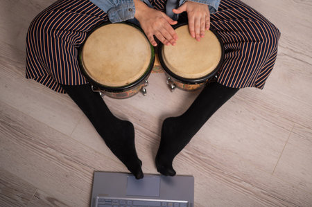 A faceless woman sits on the floor at home and watches educational videos of playing mini bongs. Girl remotely learns to play traditional ethnic drums.の写真素材