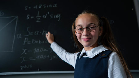 Portrait of a caucasian girl in glasses in the classroom. The schoolgirl writes the formula with chalk on the blackboard and looks into the camera.の写真素材