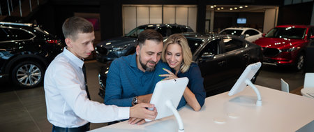 Caucasian married couple chooses a car in a car dealership on digital tablets.