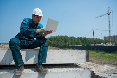 Caucasian male builder in hardhat sits on floor slabs and uses laptop at construction site.