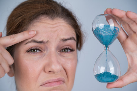 Unhappy caucasian red-haired woman looks at the hourglass and points to the wrinkles on her...